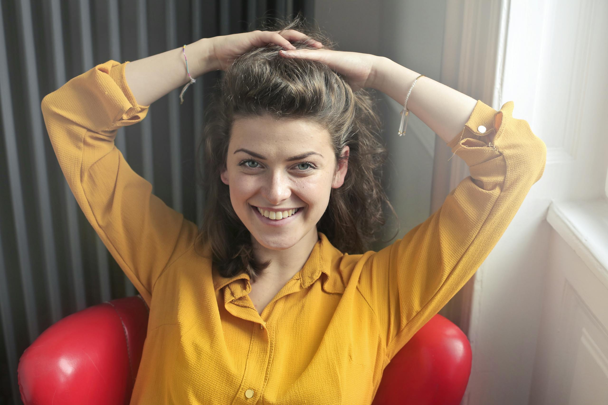 Cheerful woman sitting indoors, smiling brightly with hands behind head, wearing a yellow shirt.