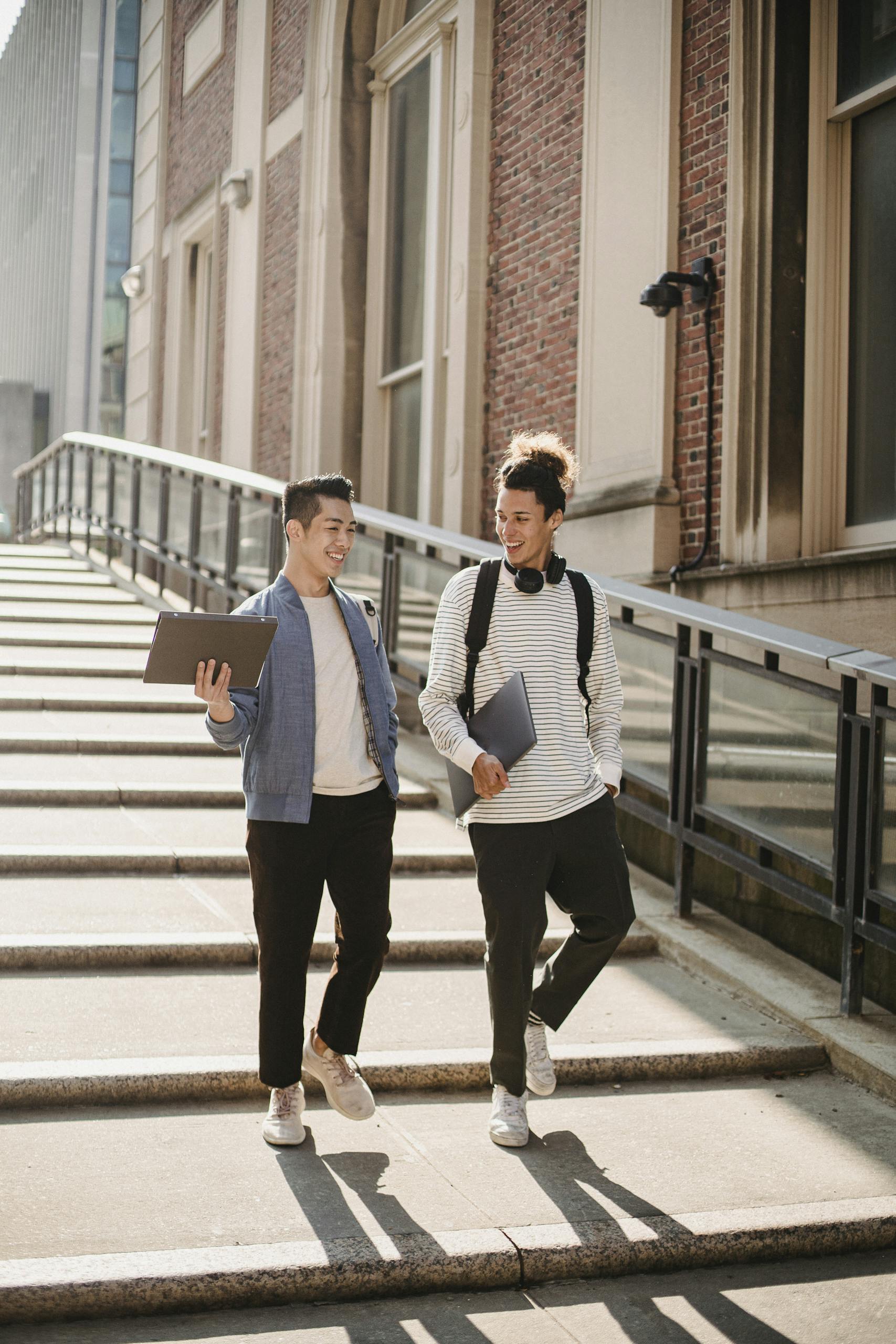 Full body of cheerful multiracial classmates with folder and laptop walking down staircase near university building