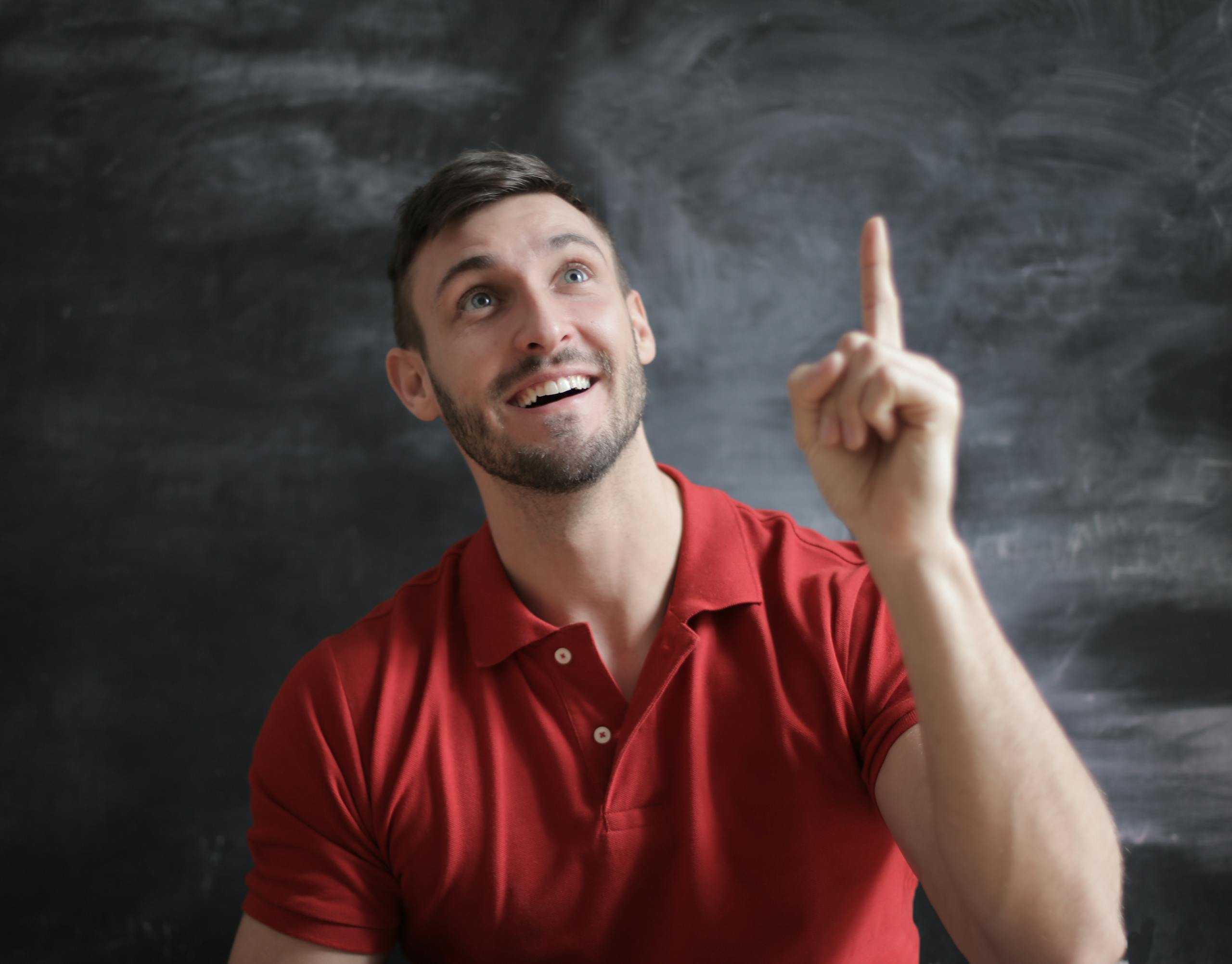 Happy young man in a red polo shirt smiling and pointing upwards in front of a blackboard.